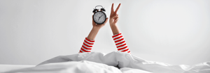 Zwei Arme ragen aus einem weißen Bett, eine Hand hält einen Wecker, die andere zeigt das Victory-Zeichen – Symbol für guten Schlaf, Erholung und Energie im Rahmen der DATAGROUP Gesundheitswoche.