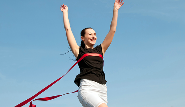 Frau läuft mit erhobenen Armen durch ein Zielband – Symbol für Motivation und Fitness.