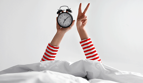 Zwei Arme ragen aus dem Bett, die eine Hand hält einen Wecker, die andere zeigt das Peace-Zeichen – Motiv für Schlaf und Erholung.
