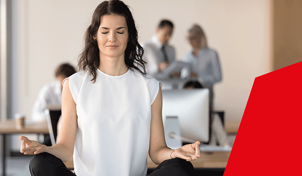 Frau sitzt im Schneidersitz im Büro und meditiert, im Hintergrund arbeiten Kollegen – Darstellung von Yoga am Arbeitsplatz.