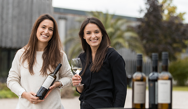 Zwei Frauen stehen draußen nebeneinander, eine hält eine Weinflasche, die andere ein Glas Weißwein in der Hand, im Vordergrund weitere Weinflaschen auf einem Tisch – Szene des DATAGROUP „Let’s Grill Together“-Events.