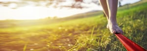 Barfuß balanciert eine Person im Sonnenschein auf einem roten Slackline-Band, das über eine grüne Wiese gespannt ist.
