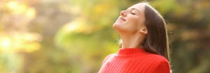 Eine junge Frau in rotem Pullover steht im Freien, schließt die Augen und atmet tief ein, während sie die warme Herbstsonne genießt.