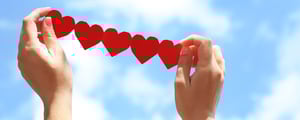Zwei Hände halten eine Girlande aus roten Papierherzen vor blauem Himmel mit weißen Wolken – Symbol für Sommer, Gemeinschaft und Herzlichkeit.