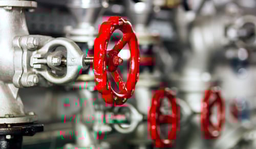 Nahaufnahme eines roten Industriehandradventils an einer Metallleitung, mit zusätzlichen Ventilen im Hintergrund, die unscharf sind.