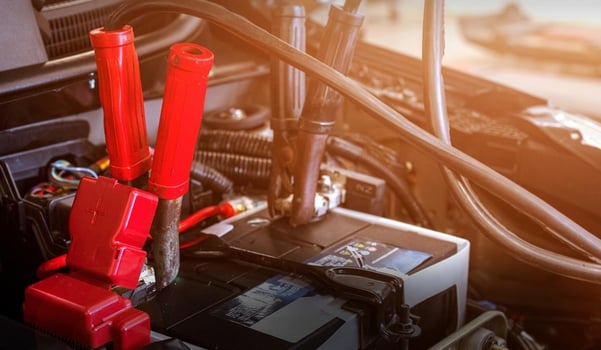 Autobatterie im Motorraum eines Fahrzeugs mit angeschlossenen roten Starthilfekabeln.