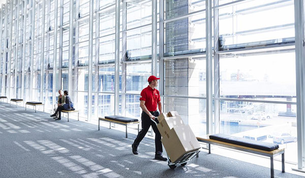 DPD-Mitarbeiter in roter Uniform schiebt einen Wagen mit Paketen durch ein helles, modernes Gebäude mit großen Glasfenstern.