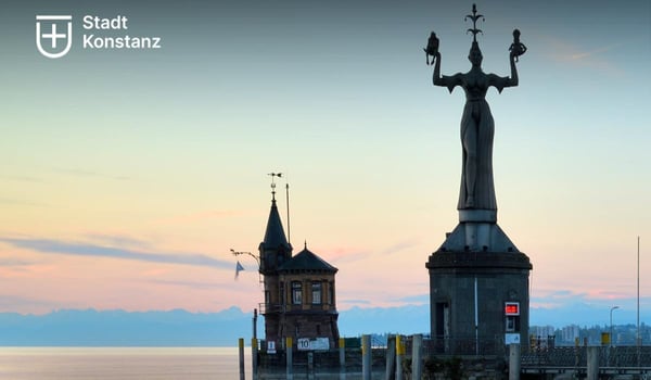 Blick auf den Hafen von Konstanz mit der Imperia‑Statue auf einem Sockel, dem historischen Hafenmeisterhaus und dem Bodensee im Hintergrund. Oben links ist das Logo der Stadt Konstanz zu sehen.