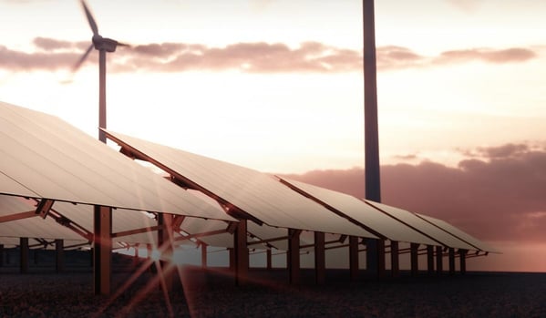Solarmodule in Reihen auf einem Feld bei tiefstehender Sonne; im Hintergrund sind Windkraftanlagen vor einem wolkigen Himmel zu sehen.