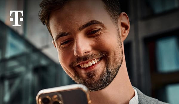 Nahaufnahme einer Person, die ein Smartphone in der Hand hält, in einer modernen Büro‑ oder Arbeitsumgebung. Oben links ist das Telekom‑Logo zu sehen.