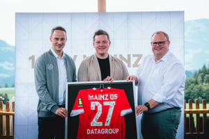 Drei Personen präsentieren ein gerahmtes, signiertes Trikot des 1. FSV Mainz 05 mit der Rückennummer 77.