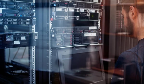 Person arbeitet an einem aufgeklappten Laptop vor einem Serverschrank mit mehreren Hardware‑Racks; links oben steht das Logo der BITBW.