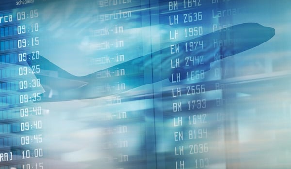Anzeigetafel eines Flughafens mit zahlreichen Flugnummern und Abflugzeiten in Blau; dahinter die schemenhafte Silhouette eines startenden Flugzeugs.