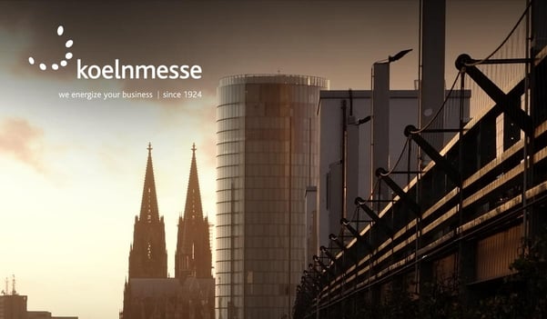 Blick auf die Kölner Innenstadt bei Sonnenuntergang mit dem Kölner Dom und einem modernen Hochhaus; links oben das Logo der Koelnmesse.