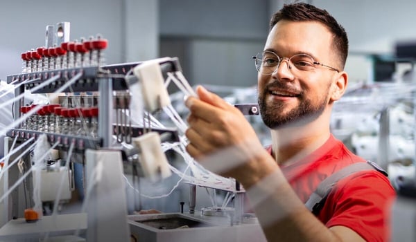 Person arbeitet an einer industriellen Maschine mit zahlreichen Fäden und Spulen; im Hintergrund ist eine Produktionshalle zu sehen. Oben links steht das Logo der MEZ GmbH.