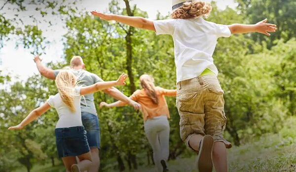 Eine vierköpfige Familie rennt fröhlich mit weit ausgestreckten Armen durch die Natur, die Eltern laufen voraus, dicht gefolgt von ihrem Sohn und ihrer Tochter.