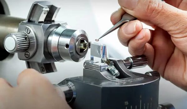 Detailaufnahme einer technischen Labor- oder Fertigungsarbeit: Eine Hand führt eine Pinzette an ein feinmechanisches Bauteil, das in einer metallischen Halterung fixiert ist. Das Equipment besteht aus präzisen Schraub- und Spannelementen sowie optischen oder mechanischen Bauteilen.