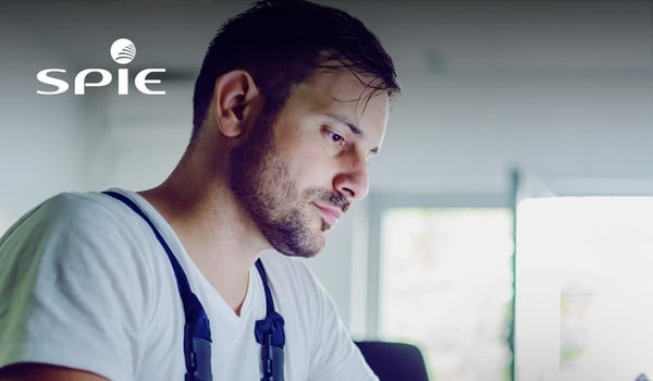 Eine Person arbeitet an einem Schreibtisch und bedient ein Tablet, während im Hintergrund ein heller Büroraum mit großen Fenstern zu sehen ist. Die Person trägt Arbeitskleidung mit Trägern. Oben links befindet sich das SPIE‑Logo.