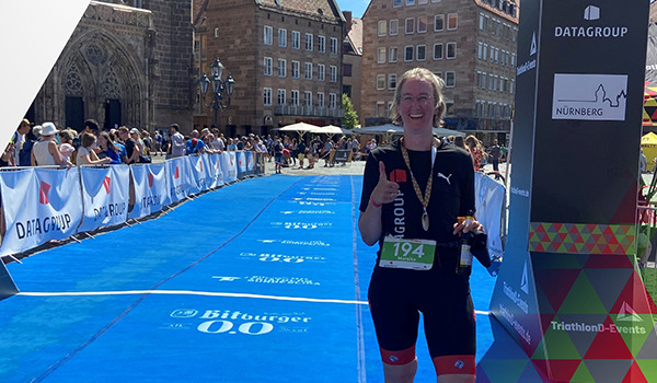 Mareike Glanz, ITSM Consultant und Risk Manager bei DATAGROUP, im DATAGROUP Triathlon-Outfit steht nach dem Zieleinlauf auf der blauen Strecke, trägt ihre Medaille und zeigt den Daumen hoch.
