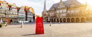 Historische Gebäude am Bremer Marktplatz bei sonnigem Wetter, im Vordergrund ein rotes grafisches Element mit stilisierter Stadtansicht.