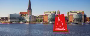 Blick über die Warnow auf moderne und historische Gebäude der Rostocker Innenstadt. Im Vordergrund ist Wasser zu sehen, im Hintergrund eine urbane Uferbebauung. Rechts im Bild steht ein rotes Segel‑Icon.