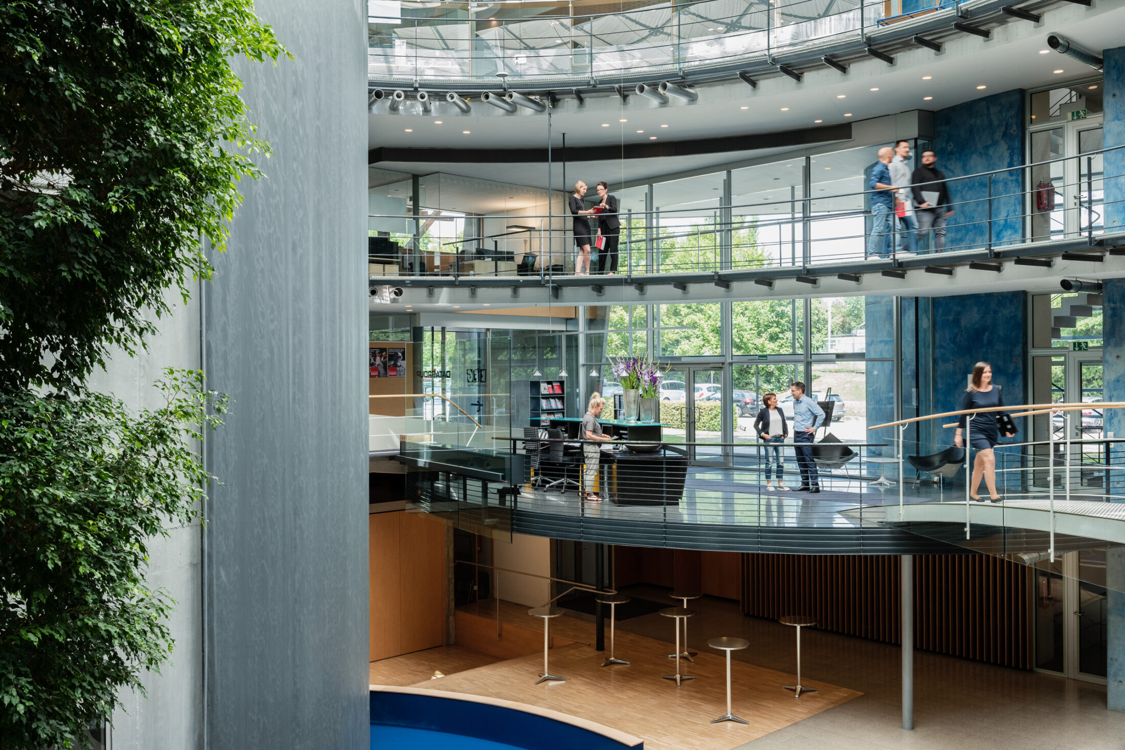 Interior view of the light-filled DATAGROUP building in Pliezhausen with several floors, a glass facade, and employees chatting in the open atrium.