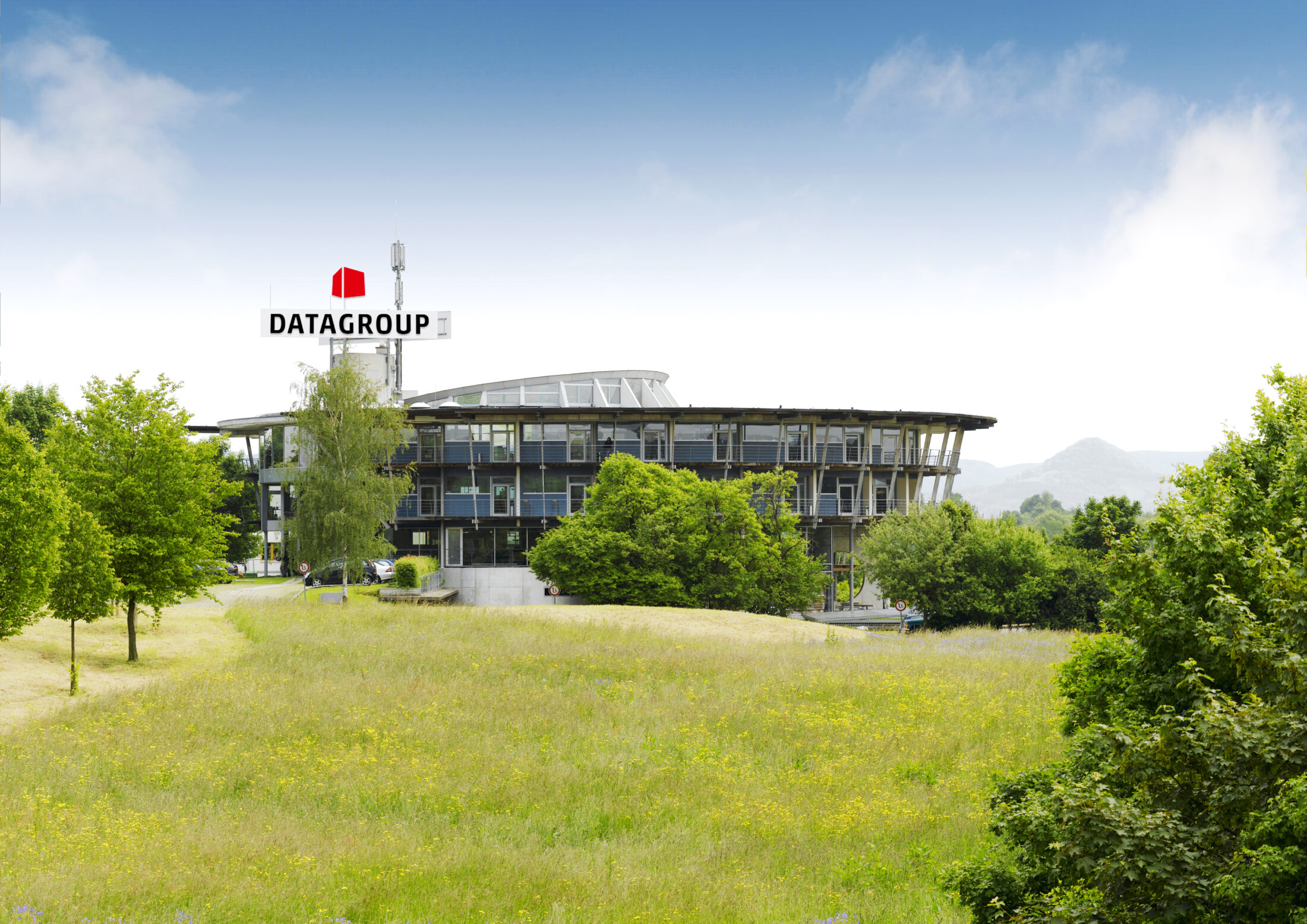 DATAGROUP headquarters in Pliezhausen, a modern office building with a circular design and a large logo on the roof, surrounded by green fields and trees.