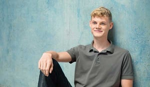 Porträt eines jungen Mannes mit blondem Haar, der ein graues Poloshirt trägt und lächelnd gegen eine blaugrüne Wand lehnt