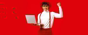 Lächelnder junger Mann in weißem Rollkragenpullover, roten Hosenträgern und Hut hält einen Laptop in der Hand und hebt jubelnd die Faust vor rotem Hintergrund.