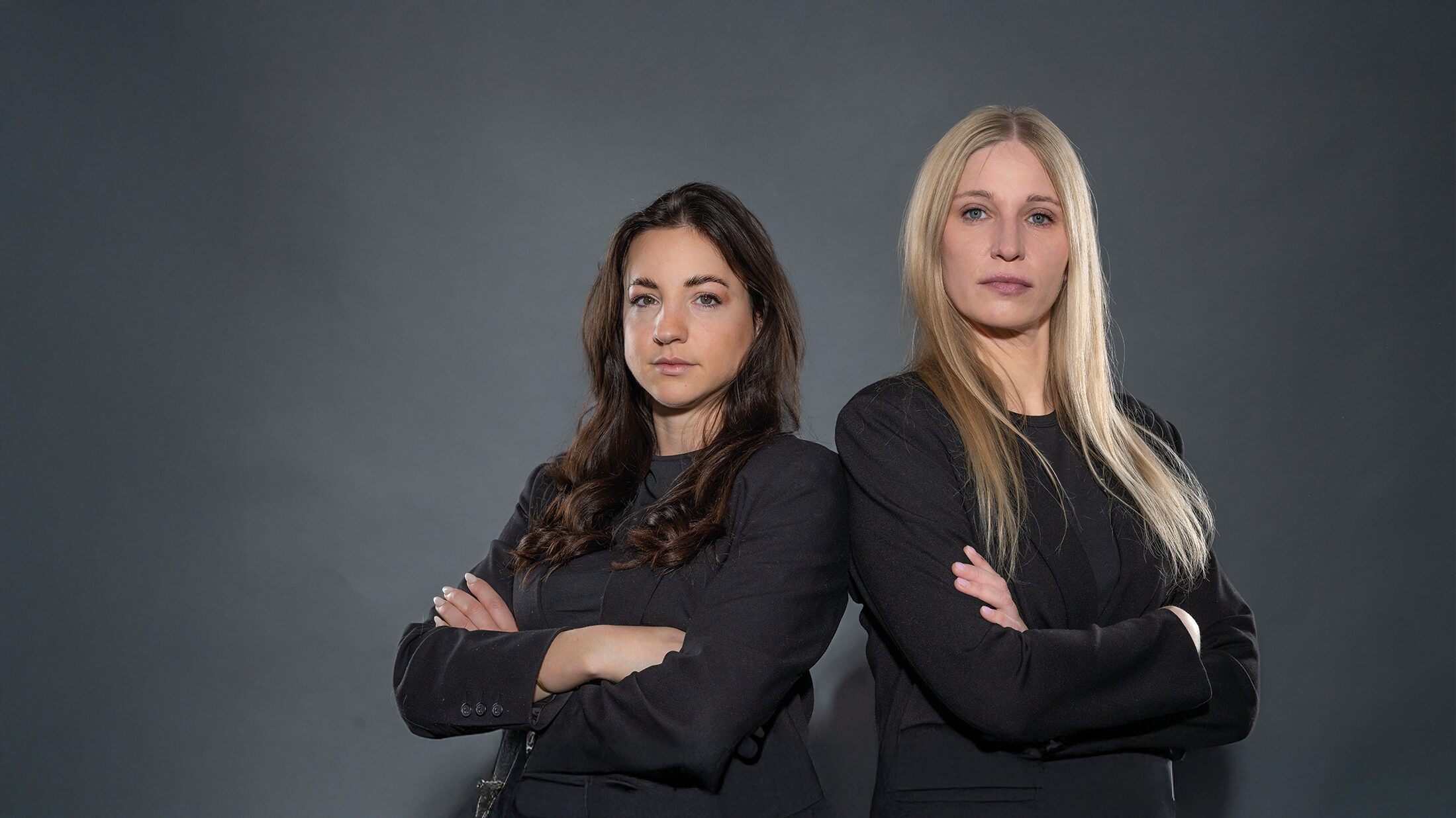 Zwei Frauen in schwarzen Outfits stehen mit verschränkten Armen nebeneinander und blicken ernst in die Kamera, vor grauem Hintergrund. Bild für den DATAGROUP Podcast Unboxing IT.