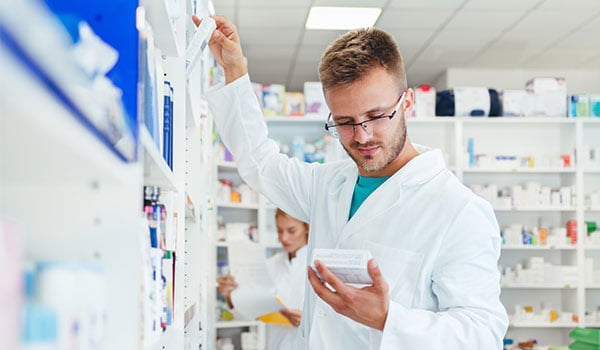 Apotheker im weißen Kittel steht in einer Apotheke und prüft die Angaben auf einer Medikamentenpackung, im Hintergrund weitere Mitarbeiterin vor Regalen mit Arzneimitteln.