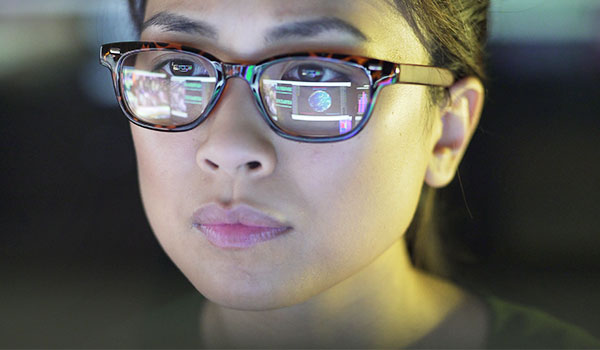 Nahaufnahme einer Frau mit Brille, in deren Gläsern sich ein Computerbildschirm mit Daten und einer Weltkugel spiegelt. Sie blickt konzentriert und fokussiert nach vorne.