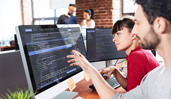 Zwei Softwareentwickler sitzen vor großen Monitoren mit angezeigtem Programmcode. Eine Frau im roten Pullover tippt auf der Tastatur, während ein Mann neben ihr auf den Bildschirm zeigt und etwas erklärt. Im Hintergrund sind weitere Personen in einem modernen Büro mit Backsteinwand zu sehen.