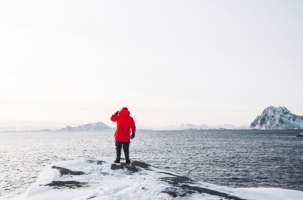 Eine Person in roter Winterjacke steht auf einem verschneiten Felsen am Meer und blickt in die Ferne über das eisige Wasser. Im Hintergrund sind verschneite Berge und eine weite, helle Winterlandschaft zu sehen.