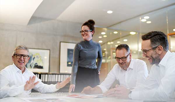 Drei Männer und eine Frau im Büro lachen und sprechen gemeinsam an einem Tisch mit Dokumenten.