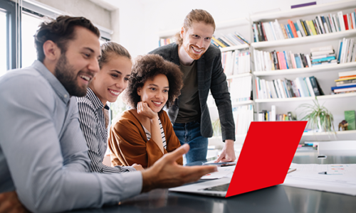 Gruppe von vier jungen Menschen sitzt zusammen in einem modernen Büro und schaut lachend auf einen roten Laptop-Bildschirm.