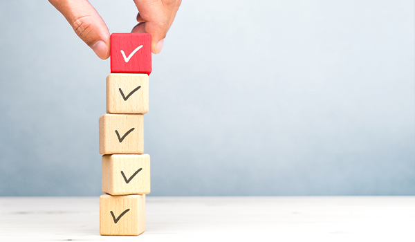 Eine Hand platziert einen roten Holzwürfel mit Häkchen auf einen Stapel von vier regulären Holzwürfeln mit Häkchen