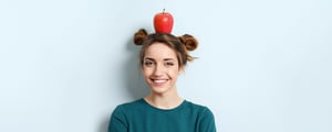 Eine junge Frau lächelt in die Kamera und balanciert einen roten Apfel auf dem Kopf – symbolisches Motiv der DATAGROUP Gesundheitswochen.