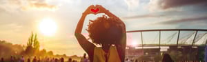Frau formt mit den Händen ein Herz in den Sonnenuntergang, im Hintergrund ein Stadion und eine Festivalstimmung mit Menschenmenge beim DATAGROUP Sommerfest 2025.