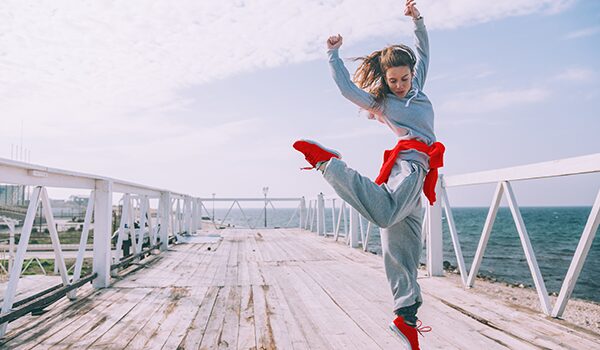 Frau springt energiegeladen auf einem Steg am Meer – Motiv für Bewegung und Firmenfitness.