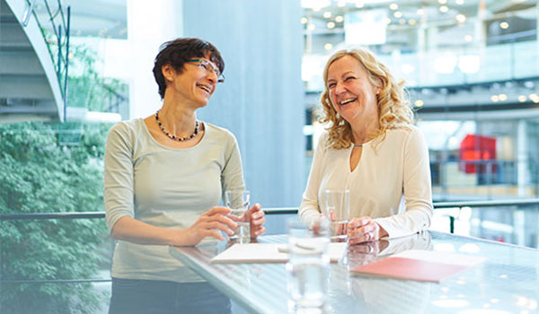 Zwei Frauen sitzen an einem Tisch in moderner Büroumgebung, lachen und unterhalten sich bei einem Glas Wasser.