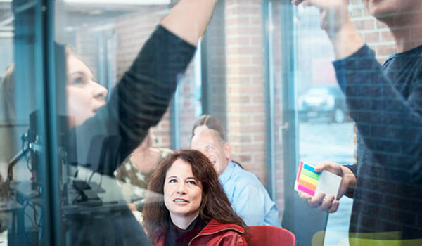 Teammeeting in einem Büro mit Glaswand, Personen schreiben auf eine Tafel mit bunten Haftnotizen, eine Frau schaut aufmerksam zu