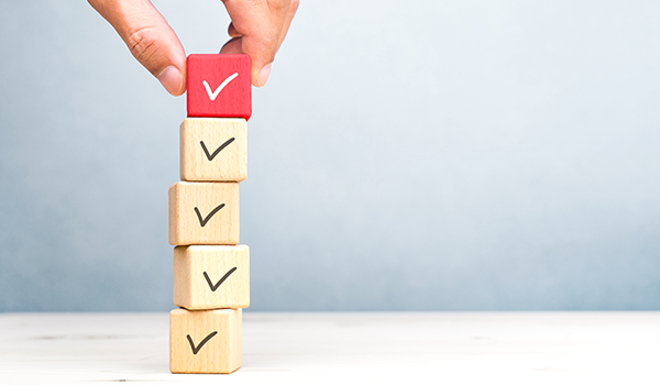 Hand stapelt Holzklötze mit Häkchen-Symbolen, der oberste Würfel ist rot hervorgehoben.