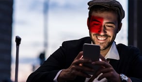 Lächelnder Mann mit Mütze und dunklem Anzug blickt auf sein Smartphone, die Reflektion eines roten Rechtecks fällt auf sein Gesicht.
