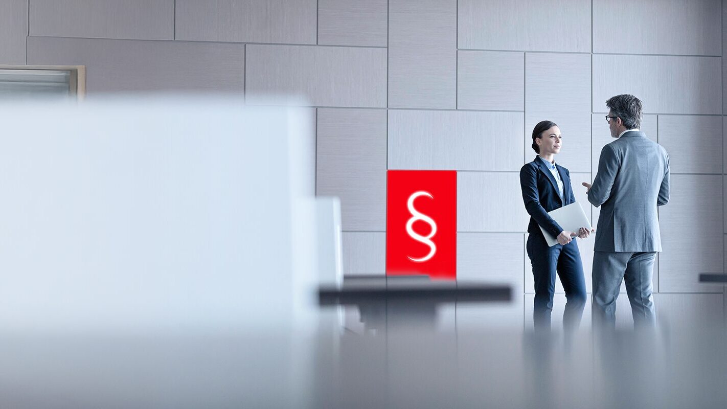 Zwei Geschäftsleute im Gespräch vor einer Wand mit rotem Paragrafen-Symbol – Sinnbild für Compliance-Services und rechtliche Sicherheit in der DATAGROUP Defense Cloud.