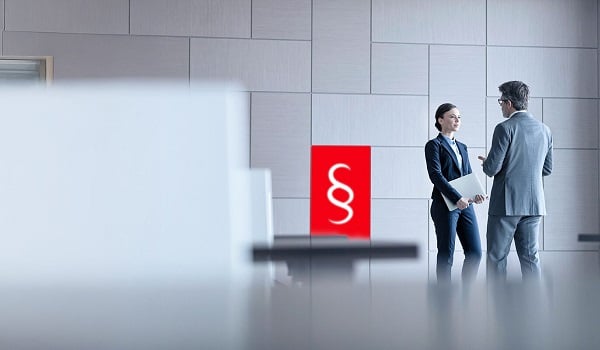 Zwei Geschäftsleute im Anzug unterhalten sich in einem modernen Bürogebäude, im Vordergrund ist ein rotes Symbol mit weißem Paragrafenzeichen zu sehen.