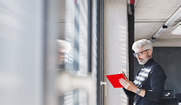 Mann mit grauen Haaren und Brille steht am Fenster in einem modernen Büro und arbeitet konzentriert auf einem roten Tablet.