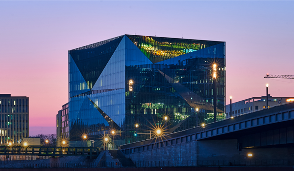 Moderne Glasfassade des Drees &amp; Sommer Gebäudes bei Sonnenuntergang, mit leuchtenden Lichtern und Spiegelungen in der Fassade.