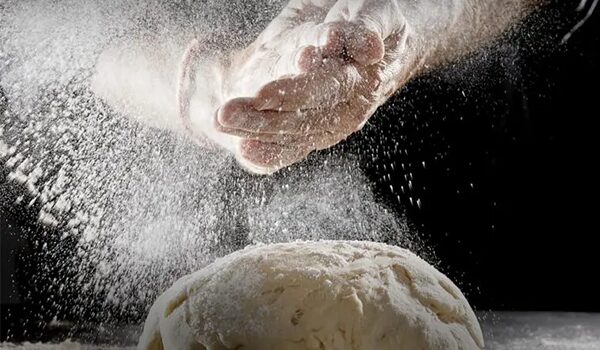 Nahaufnahme von Händen, die Mehl über Teig auf einer dunklen Oberfläche streuen, wobei Mehlpartikel während der Backvorbereitung in der Luft schweben.
