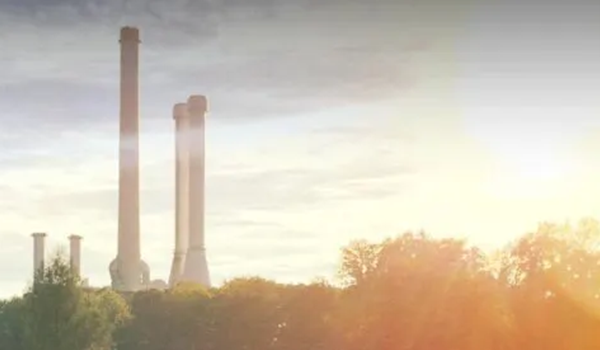 Bild von Industrieschornsteinen, die bei Sonnenaufgang über den Baumwipfeln aufragen, während warmes Sonnenlicht die Szene beleuchtet.